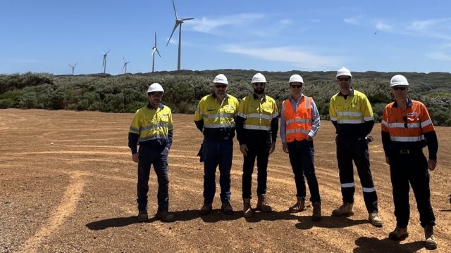 Six men in hi-viz clothing and white hard hats standing in a line on a dusty area located in front of five onshore wind turbines. The sun is shining from a clear blue sky.