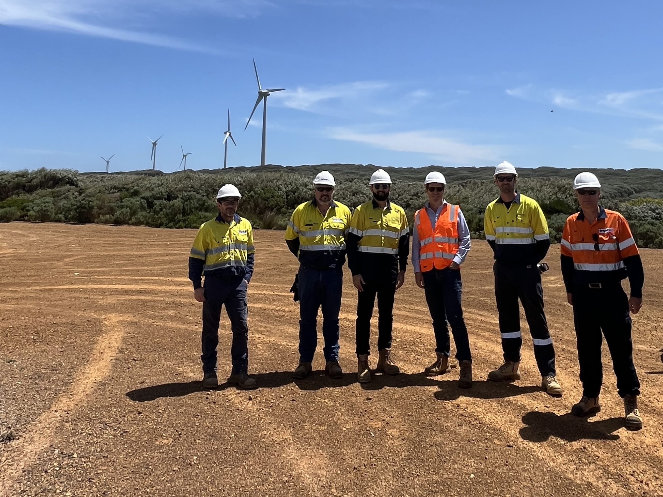Six men in hi-viz clothing and white hard hats standing in a line on a dusty area located in front of five onshore wind turbines. The sun is shining from a clear blue sky.