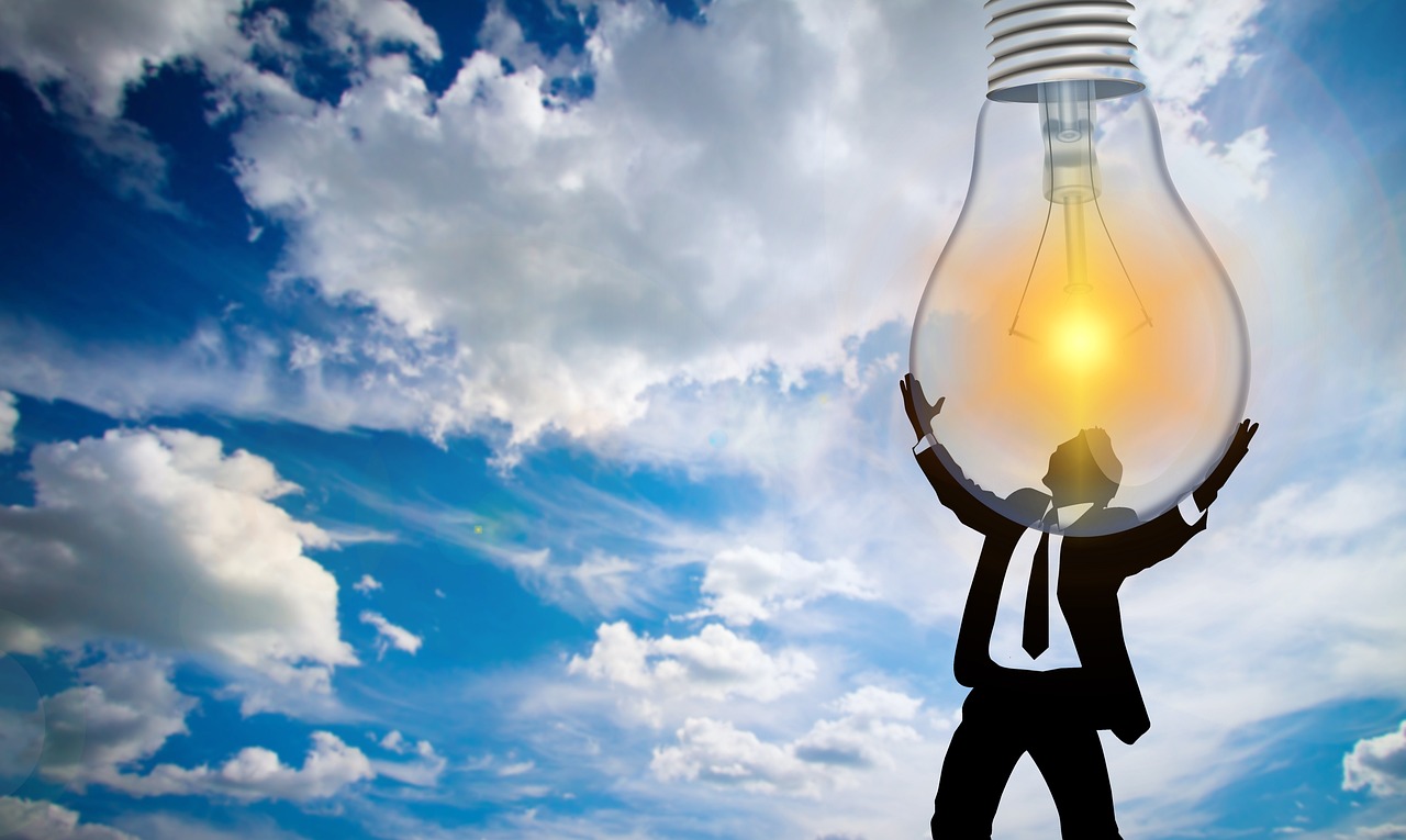 Person holing a light bulb lit by the sun. In the background there are white clouds on a blue sky